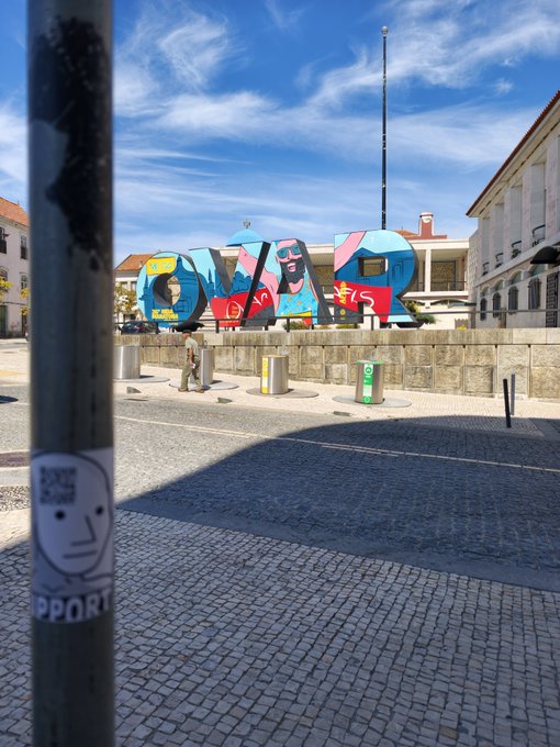 A sunny street view features a large colorful sign spelling 'AVEIRO' decorated with stylized portraits, viewed past a lamppost bearing a small sticker.