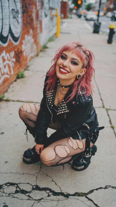 The image features a woman with pink hair, wearing a leather jacket and fishnet stockings while squatting down on the sidewalk. She has a smile on her face as she poses for the picture. The caption of the photo includes various social media handles such as @stringerjohn007, @laf131, and @delerat7, which are likely used to share or promote the image.