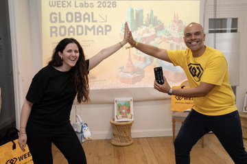 The image features two people standing next to each other, both holding up their cell phones with big smiles on their faces. They are posing for a photo at an event where they are celebrating the global road map. The background of the picture includes a large screen displaying the text "WEEX x Pudgy Penguins at #ProofOfTalk was a blast! @pudgypenguins BIG congrats to @rose_garden5 - onsite AI Trading Comp champ AI trading trading comp champ AI trading trading comp".