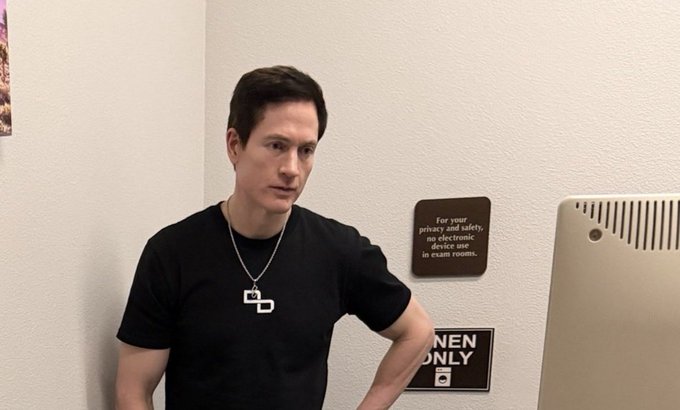 A photo of a man in a black t-shirt and chain standing next to a restroom sign and an iMac computer.
