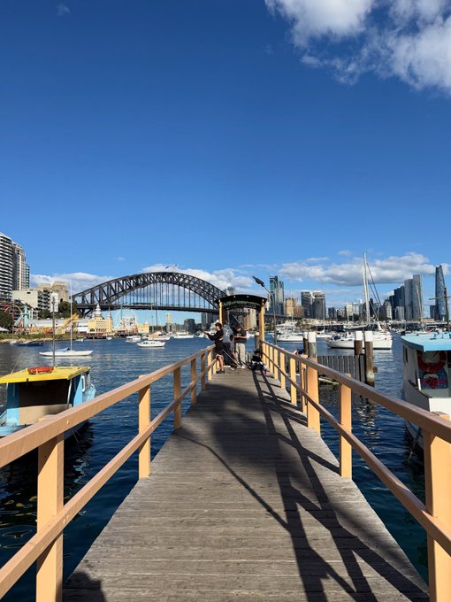 A wooden pier with yellow railings extends toward a harbor filled with boats, leading toward the Sydney Harbour Bridge and city skyline under a clear blue sky.