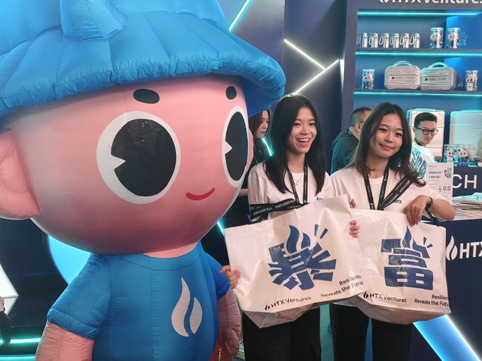 Two women smiling and holding HTX Ventures tote bags alongside a large pink and blue mascot at an event booth.