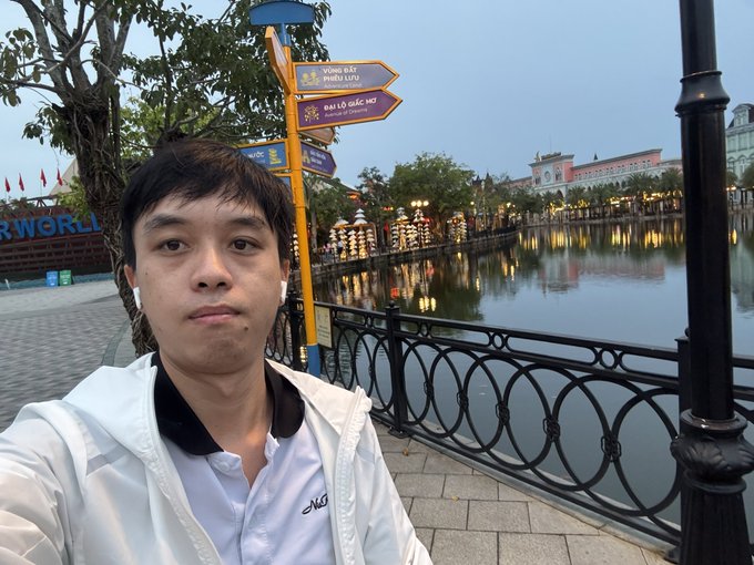 A young man taking a selfie by a lake at dusk, wearing a white jacket, with directional signs in Vietnamese pointing to 'VƯỜN ĐT PIETRO LURU' and 'ĐẠI LỘ ĐẶC MÔI' visible in the background.