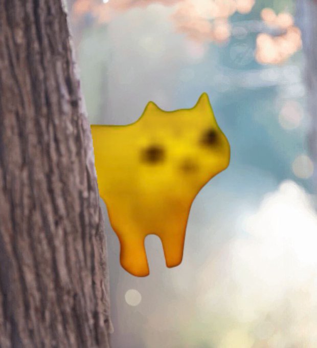 A cute yellow cat-shaped squishy or charm with a simple smiling face hanging against a blurred background with tree bark visible on the left side.