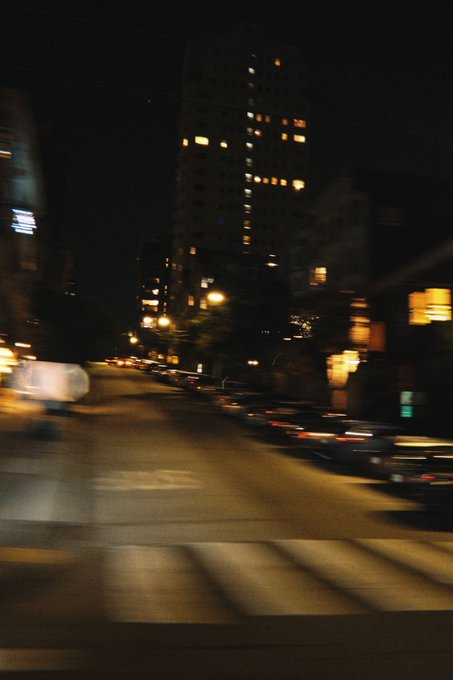A blurry nighttime city street view showing illuminated building windows and parked cars along the road.