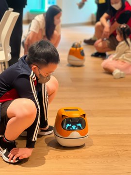 A young boy crouches down to face a small, orange and black robot on a wooden floor, while other children, parents, and caregivers gather around in the background at Ronald McDonald House Charities Hong Kong. The scene reflects the post's message of Floki Minibots bringing joy to families in a caring environment.