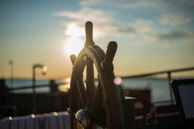 A serene sunset photograph featuring a ship's wooden steering wheel in silhouette against a glowing sky, with the sun creating lens flare over what appears to be a harbor or waterfront.