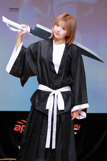 A photograph of a woman in cosplay as a Bleach anime character (likely Rukia), wearing a black shihakusho with a white obi and carrying a large sword on her shoulder, taken at a convention event.