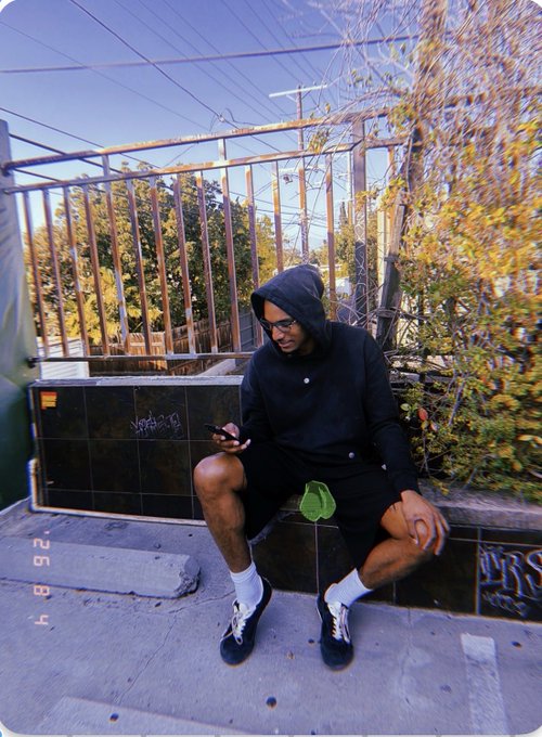 A person wearing a black hoodie, black shorts, glasses, and a beanie sits on a tiled ledge checking their phone, with power lines and trees in the background.
