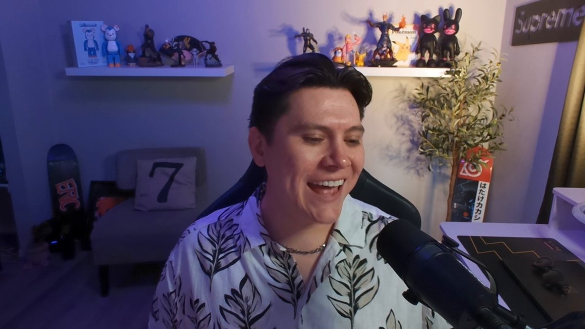 The image features a man sitting behind a microphone, smiling at the camera while wearing a white shirt with black leaves printed on it. The background includes various figurines and toys, suggesting that this scene takes place in a room filled with collectibles or possibly an office dedicated to video game merchandise.