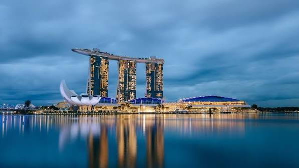 A twilight photograph of the Marina Bay Sands resort in Singapore, with its three towers topped by the SkyPark and the lotus-shaped ArtScience Museum visible, reflecting in the calm water.