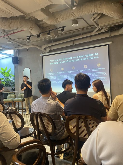 A photo of an indoor presentation or seminar where a speaker stands in front of a projection screen displaying Vietnamese business content about AI solutions, while an audience of young adults sits in chairs facing the presentation.