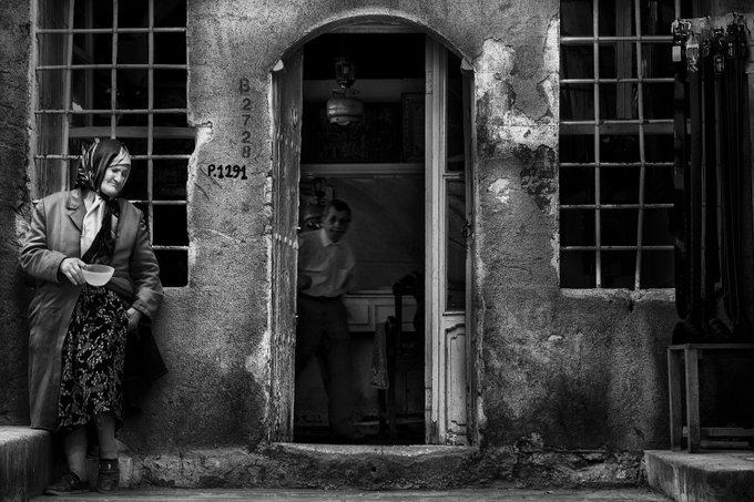 The image shows a woman sitting outside of an old building with a doorway that has been opened up. She appears to be wearing a coat and holding a cup. The door also has a sign on it, which reads "BZT". In the background, there is another person standing near the doorway.