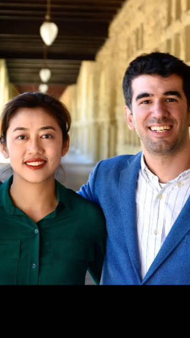 A smiling young Asian woman in a green blouse stands beside a smiling young man in a blue blazer in a warmly lit corridor, with no visible text. The image relates to the post by visually representing the human collaborators and creators at the heart of Pi Network's evolution, with their upbeat demeanor mirroring the optimistic vision of creators transforming ideas into applications on a decentralized platform.