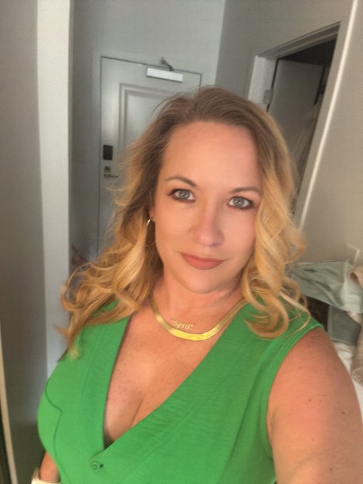 A selfie of a blonde woman with wavy hair wearing a green v-neck top and a gold choker necklace, standing in what appears to be a hotel room with a door visible behind her.