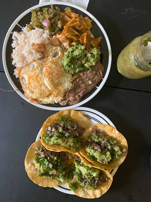 An overhead photo of a Mexican meal featuring a bowl with rice, fried eggs, guacamole, refried beans, and tortilla chips, alongside a plate of carne asada tacos topped with guacamole, with a green smoothie on the side.