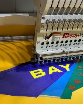 A HappyJapan embroidery machine is stitching the bold yellow "BAYC" lettering onto a vibrant purple, yellow, and blue burgee flag, showcasing the production process for the Bored Ape Yacht Club raffle giveaway where fans can sign up to win one of these flags by August 5th, 2026.
