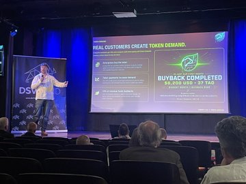 A presenter speaks on stage at the DSV Fund event in London, with a large screen displaying "REAL CUSTOMERS CREATE TOKEN DEMAND" and "BUYBACK COMPLETED: $6,800 USD - 97 TAO," highlighting DSV's recent buyback achievement for the Bittensor ecosystem.
