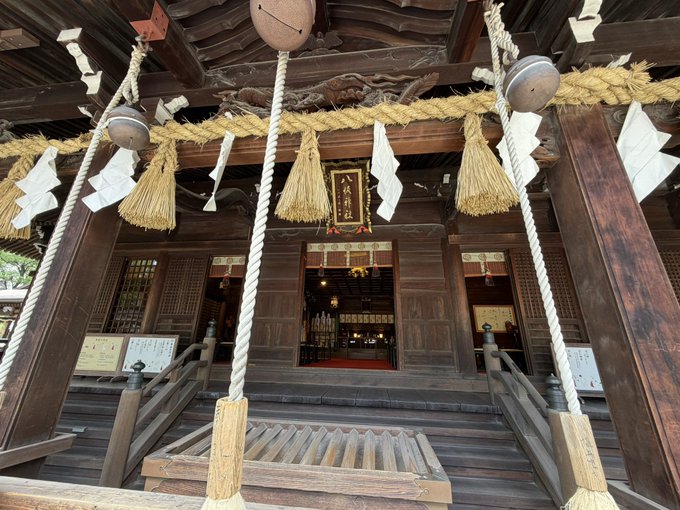 The entrance of a traditional Japanese Shinto shrine with a large shimenawa rope, shide paper streamers, and a wooden plaque reading 八坂神社 (Yasaka Shrine).