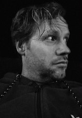A black-and-white close-up portrait shows a bearded man with tousled hair looking off to the side against a dark background.