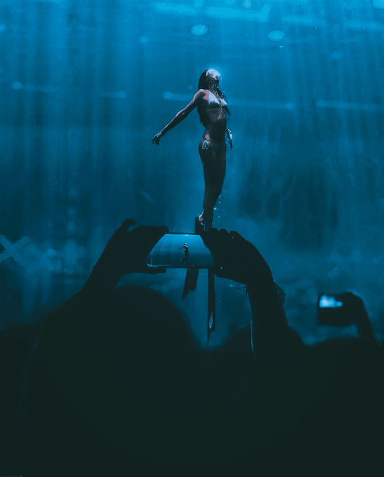 An underwater photograph shows a performer standing gracefully atop a submerged shark tank while audience members in silhouette watch and take photos with their phones.