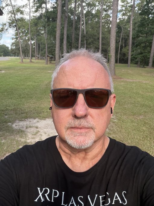 A man takes a selfie outdoors in a grassy park with trees in the background, wearing dark sunglasses and a black t-shirt that reads "XRP LAS VEGAS."
