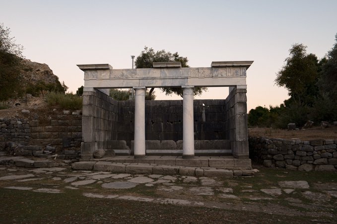 Ancient stone ruins featuring a colonnade with white pillars and entablature stand at dusk, surrounded by stone walls and trees in a historical archaeological site.