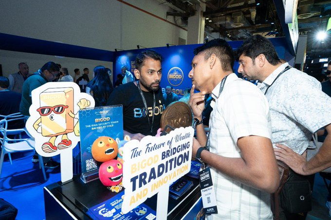 The image shows a group of people gathered around a table at an event, where they are looking at various items displayed for sale. The table features several items such as a sign that says "The Future of Music Borrowing Tradition", which is likely related to music streaming services or digital downloads. There are also two men standing near the table, one wearing sunglasses and another with a lanyard around his neck. A man in a white shirt can be seen talking to someone else at the event. The atmosphere appears to be casual and social as people engage in conversation and browse through the items on display.