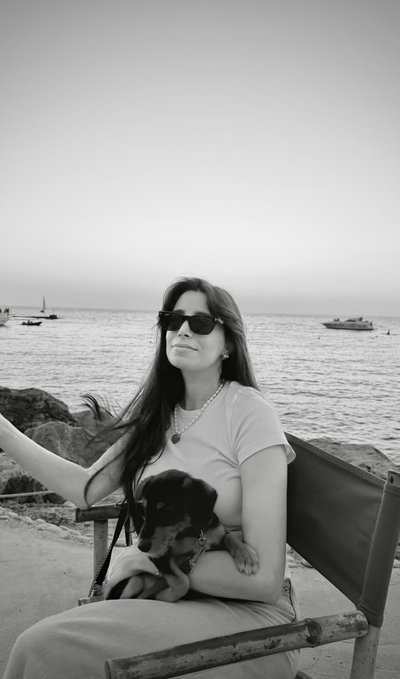 A black-and-white photo of a woman wearing sunglasses sitting on a bench by the sea, holding a small black puppy in her lap.