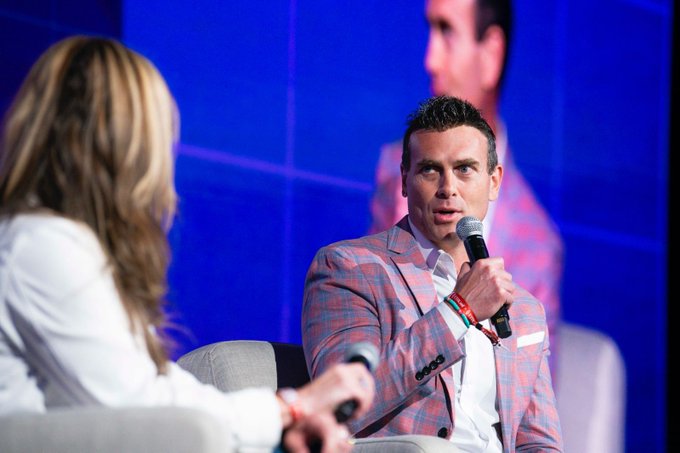 The image shows a man wearing a pink suit talking into a microphone while sitting at a table or stage. The man appears to be engaged in a conversation or presentation with another person who is also present in the scene, possibly an audience member.