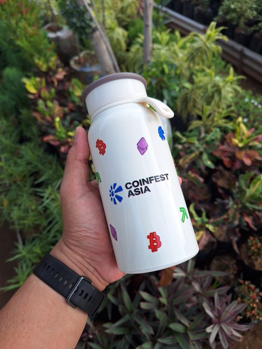 A hand holds a white tumbler branded with "COINFEST ASIA" and decorated with various cryptocurrency logos, including Bitcoin, as a freebie from the crypto event. The tumbler represents the stream of branded merchandise the poster has been receiving since getting into crypto.