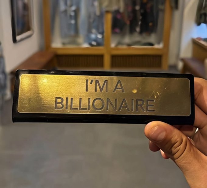 A hand proudly displays a brass plaque engraved with "I'M A BILLIONAIRE," shared with the caption "Claim this shiiii" in a joking declaration of newfound wealth. The background appears blurred, possibly a public space, emphasizing the playful boast of the shiny nameplate.