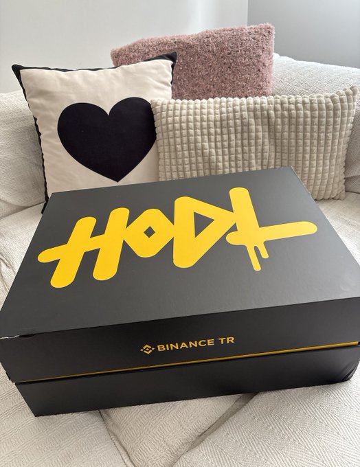 A black surprise box with bold yellow "HODL" graffiti-style text on top sits on a couch, featuring the "BINANCE TR" logo along its side. The unopened package aligns with the user's excitement about being one of the first recipients of Binance TR's gift box, which they are keeping secret from others.