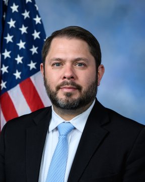 The image shows a headshot of a man in a dark suit with a light blue striped tie, standing in front of an American flag against a blue backdrop. This appears to be an official portrait of a U.S. Senator, likely one of the senators mentioned in the accompanying post about the Clarity Act, possibly Sen. Ruben Gallego.
