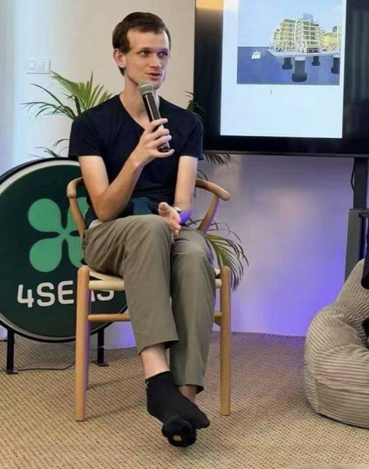 The image features a man sitting in a chair, holding a microphone and speaking into it. He appears to be at a presentation or event where he is addressing an audience. The background includes a TV screen displaying a building, suggesting that this might be related to architecture or urban planning. There are also potted plants visible in the scene, adding some greenery to the environment.