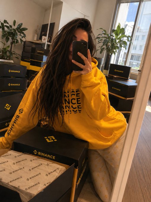 A person takes a mirror selfie with their phone while surrounded by stacks of black "BINANCE" branded boxes, preparing to send out the #BinanceConnect prize packages to the giveaway winners announced in the post.