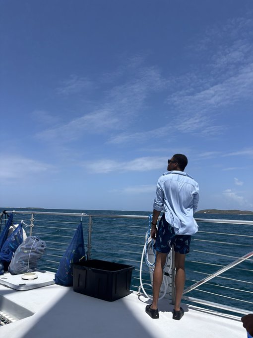 A man in a light blue shirt and floral shorts stands on a boat, looking out over blue ocean waters under a partly cloudy sky.