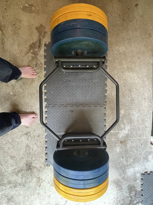 A top-down view of a hex bar (trap bar) loaded with yellow and blue weight plates resting on a diamond-plate mat, with a person's bare feet visible to the left.