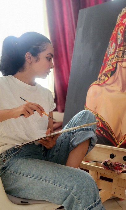 A young woman with dark hair pulled back sits in profile, holding a paintbrush and palette while working on a vibrant portrait of a figure with an ornate red and gold headdress.