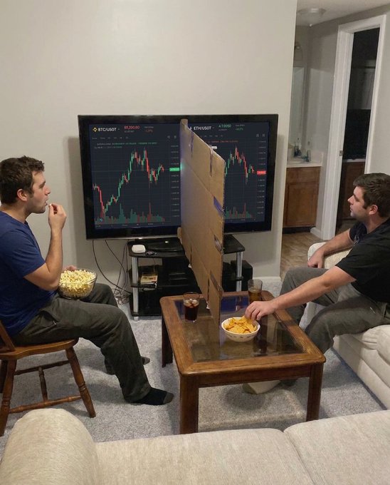 Two men sit in a living room staring intently at separate cryptocurrency charts on a dual-monitor setup, one displaying BTC/USDT at 56,200.00 and the other showing ETH/USDT at 1,100.00, with bowls of snacks and drinks on the coffee table between them. The scene perfectly captures the post's humor about being emotionally tethered to different coins on Binance, as both friends sit glued to their screens while seemingly ignoring each other in favor of monitoring their respective portfolios.