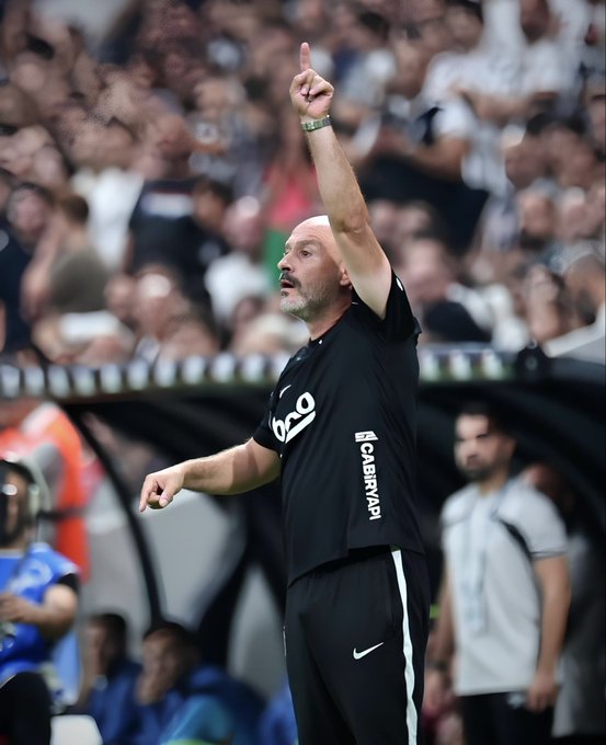 A bearded coach in a black uniform with "bta" and "CARNYAPI" sponsor logos points upward with his index finger while giving directions, with a blurred crowd visible behind him. The caption "𝑽𝒊𝒏𝒄𝒆𝒏𝒛𝒐 𝑰𝒕𝒂𝒍𝒊𝒂𝒏𝒐 Hedef belli. #UNIBJK" identifies the coach as Vincenzo Italiano, emphasizing his determined focus on a clear objective during the match.