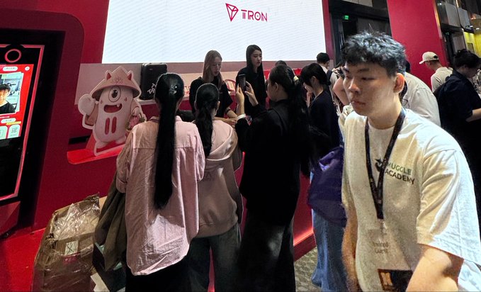 People gathered at a TRON blockchain booth at what appears to be a crypto convention, with a young man in a white 'Muscle Academy' t-shirt in the foreground.