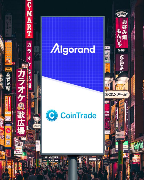 The image shows a large billboard with an advertisement for Algorand, a cryptocurrency, and a coin trading platform called CoinTrade. The billboard features a blue background with white text that reads "Algorand" and "coinTrade". Below this, there is a smaller white banner displaying the logo of CoinTrade in Japanese. The image also includes a photo of a busy street scene filled with people walking around at night.