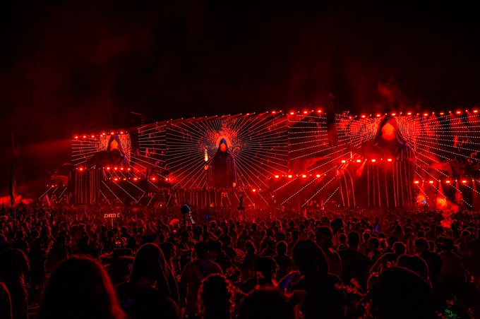 A large nighttime music festival crowd bathed in intense red lighting, with an elaborate stage featuring a sunburst design and large arched cutouts displaying a face.