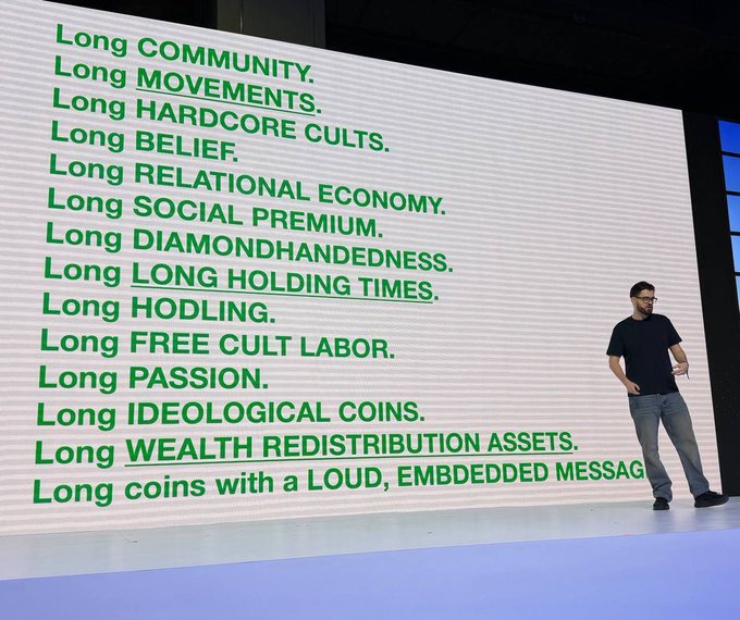 A presentation slide listing principles including 'Long COMMUNITY', 'Long MOVEMENTS', 'Long HARDCORE CULTS', 'Long BELIEF', 'Long RELATIONAL ECONOMY', 'Long DIAMONDHANDEDNESS', and 'Long WEALTH REDISTRIBUTION ASSETS', with a presenter standing to the right.