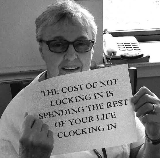 A smiling man with glasses holds up a sign reading "THE COST OF NOT LOCKING IN IS SPENDING THE REST OF YOUR LIFE CLOCKING IN," humorously emphasizing the urgency of taking action now—reinforcing the post's message about seizing opportunities to build wealth onchain before time runs out.