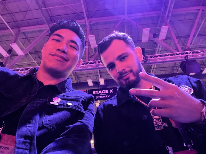 Two men pose for a selfie at a large event; a sign in the background reads 'STAGE X' and 'foundry'.