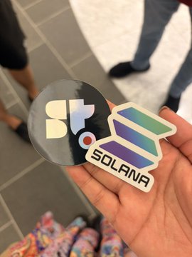 Hand holding two stickers: a black circular one with 'Sp' logo and a colorful holographic 'SOLANA' sticker, with a person's legs visible on a tiled floor in the background.