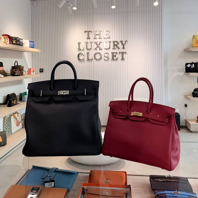 Two luxury handbags, one black Birkin-style and one burgundy Birkin-style, are displayed in 'THE LUXURY CLOSET' showroom with shelves of designer goods visible in the background.