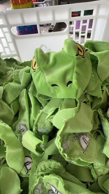 A pile of green frog-themed plush items or fabric pieces in a white basket, with one frog face visible at the top.